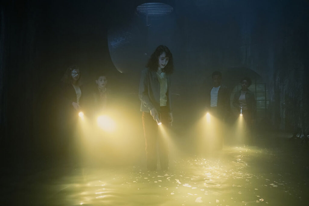 The children are standing in the water in the sewers. They look at the water, lit by their flashlights.