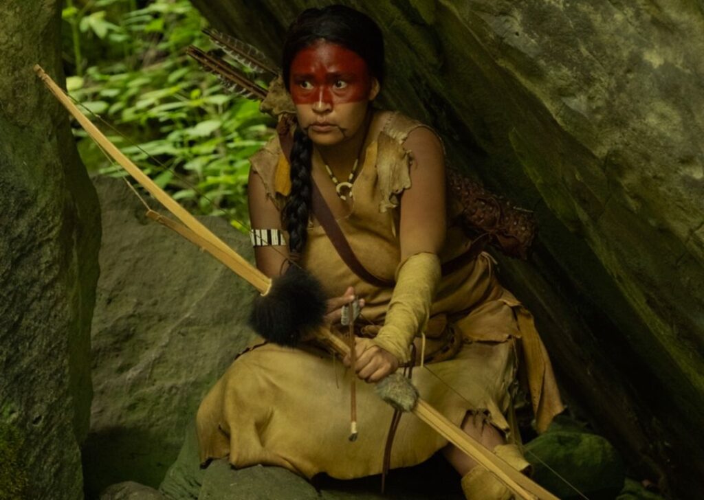 It: Welcome to Derry - an indigenous woman names Sesqui is hiding under a rock and holding a bow